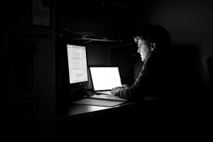 Man at a desk in a dark room, lit by two computer monitors