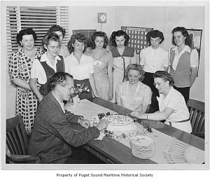 A group of women gather around a man cutting a birthday cake in a 1940s office